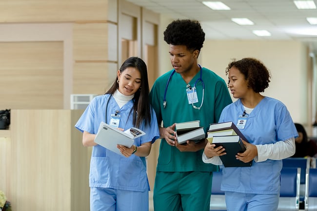 Three medical assistant students walking through a medical facility holding a stack of books.