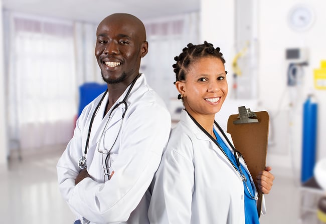 Medical assistant and doctor standing back to back smiling at the camera.