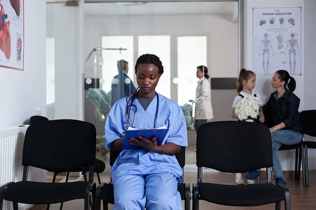 Medical assistant sitting in waiting room with a clip board writing something down - patients sitting and other medical staff moving in the background.