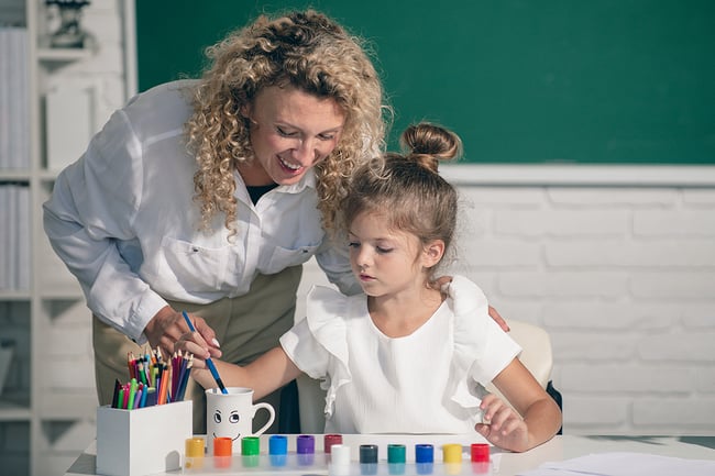 Young female teacher with curly hair leaning over young girls shoulder helping her learn how to paint.
