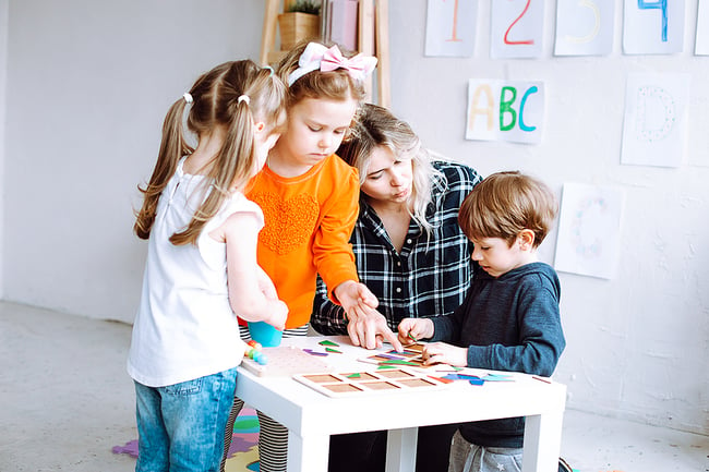 young blond female preschool teacher playing with three students in a classroom
