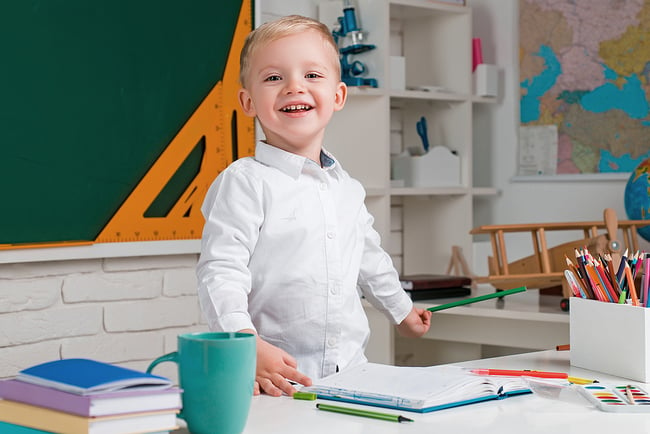 Preschool student standing on the teacher's desk chair smiling and holding a pencil.