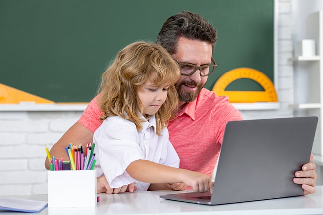 Male teacher helping preschool student use a laptop in the classroom.