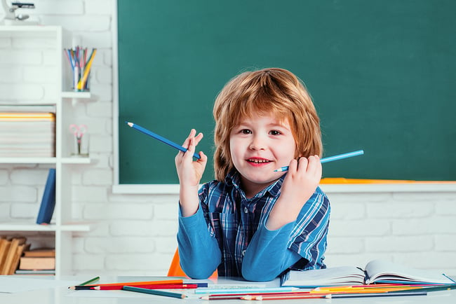 little boy sitting in a classroom with chalkboard behind him holding colored pencils