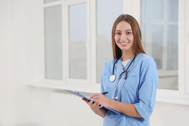 Female medical assistant wearing blue scrubs holding a clipboard and pen.
