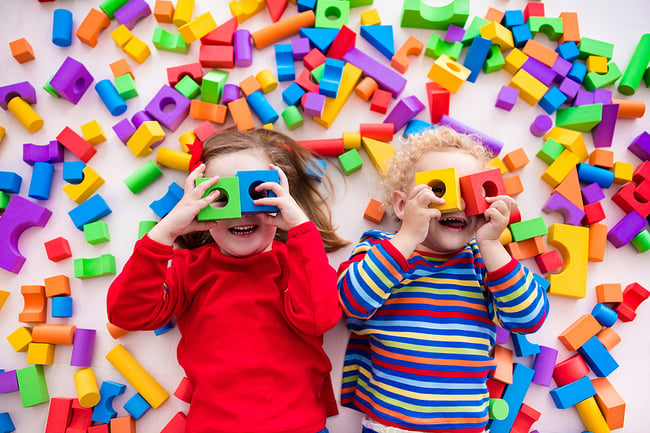 Two preschool students laying in toys peeking through blocks and smiling.