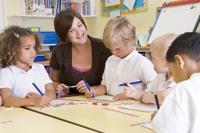 Preschool teacher sitting with some young students at a classroom table.