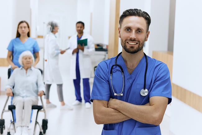 Male medical assistant standing in hallway of a hospital with other medical assistants and doctors in the background. 