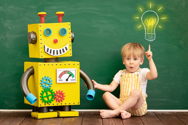 Preschooler sitting on the classroom floor in front of a green chalkboard holding hands with a colorful robot.