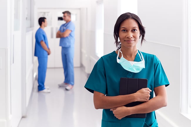 Smiling female medical assistant standing in the hallway of a medical facility with two medical assistants in the background slightly blurred.