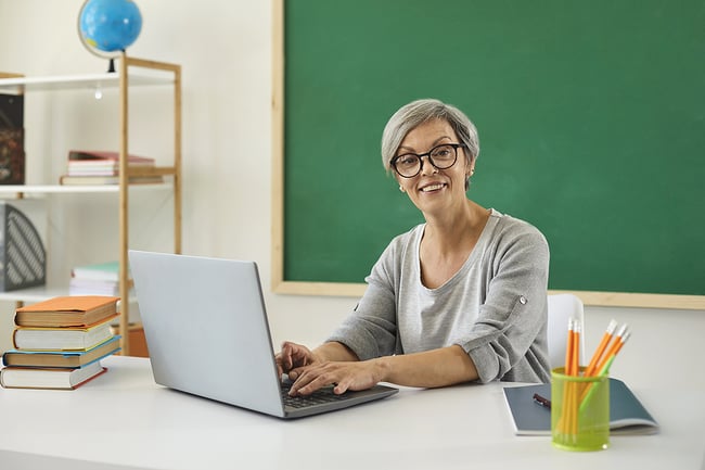 Older woman sitting at a desk in front of a chalkboard with a laptop.