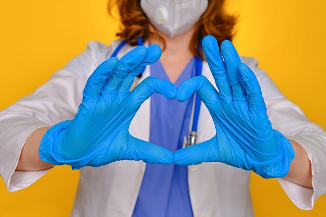 Healthcare worker wearing rubber gloves and holding hands in a heart shape toward the camera.