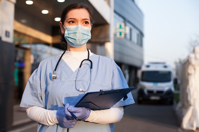 Medical assistant standing outside a hospital wearing scrubs and stethoscope holding a clipboard. 